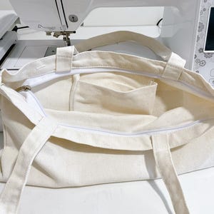 May include: Cream-colored canvas tote bag with a white zipper and two handles. The bag has a rectangular shape with a small pocket inside. The bag is open, revealing the interior. The background includes a white sewing machine.