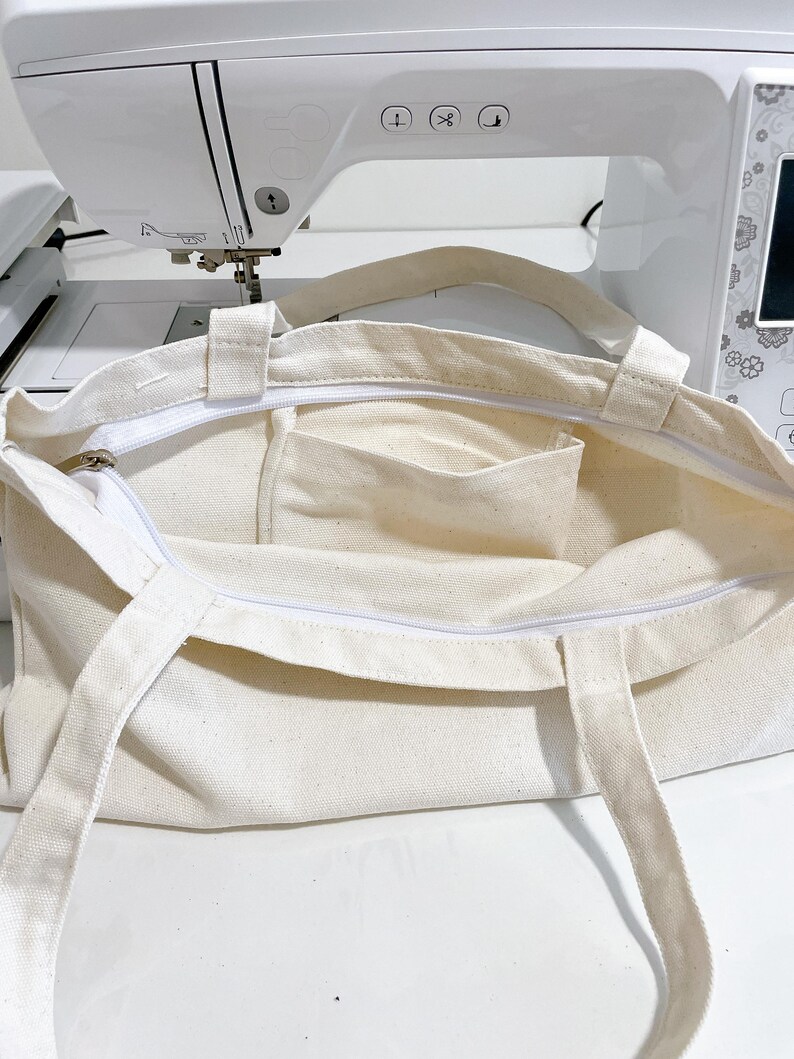 May include: Cream-colored canvas tote bag with a white zipper and two handles. The bag has a rectangular shape with a small pocket inside. The bag is open, revealing the interior. The background includes a white sewing machine.