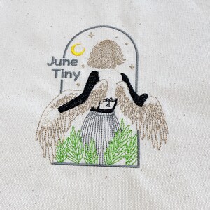 May include: Embroidered design on a canvas tote bag. The design features an angel with outstretched wings, a striped skirt, and the words "June Tiny." The angel is framed by an arch with a crescent moon and stars.