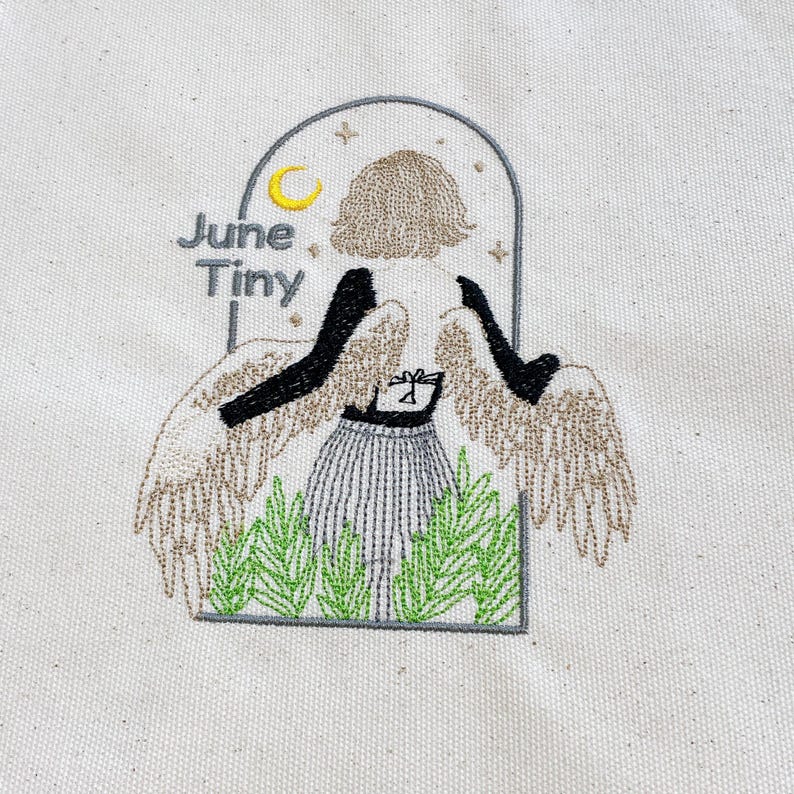 May include: Embroidered design on a canvas tote bag. The design features an angel with outstretched wings, a striped skirt, and the words "June Tiny." The angel is framed by an arch with a crescent moon and stars.