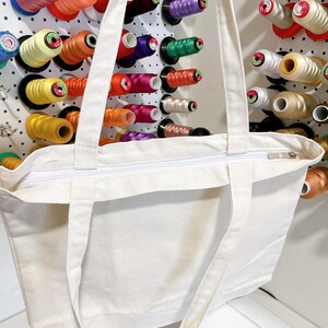 May include: A white canvas tote bag with a zipper closure and long straps. The bag is in front of a pegboard filled with colorful spools of thread in various colors, including red, orange, yellow, green, and blue.