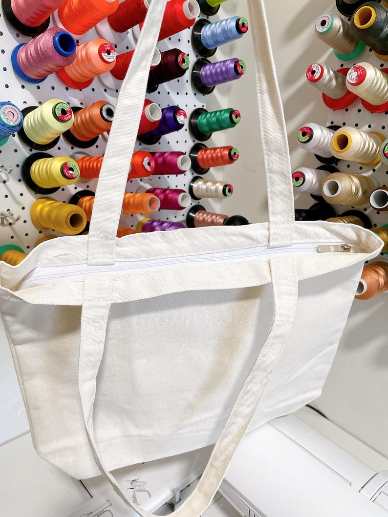 May include: A white canvas tote bag with a zipper closure and long straps. The bag is in front of a pegboard filled with colorful spools of thread in various colors, including red, orange, yellow, green, and blue.