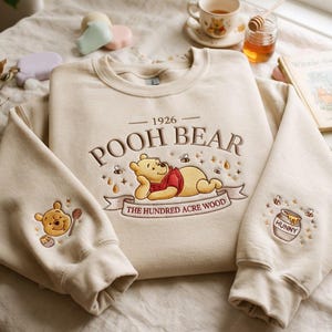 Peut inclure: Un sweat-shirt à col rond beige avec une broderie représentant Winnie l'ourson allongé, accompagnée du texte 1926 POOH BEAR THE HUNDRED ACRE WOOD. Les manches présentent des détails brodés de Winnie et d'un pot de miel étiqueté HUNNY.