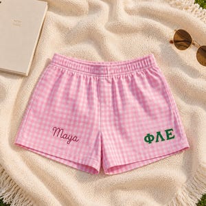 May include: Pink and white gingham shorts with an elastic waistband. The left leg features the name Maya embroidered in pink script, while the right leg displays the Greek letters ΦΛΕ embroidered in a bold, dark green block font.