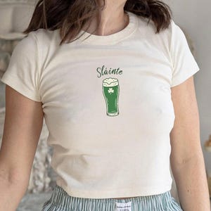 May include: Cream-colored crop top featuring the word "Sláinte" above a green beer glass with a shamrock. The shirt is a short-sleeved, crew neck style. The model is wearing striped pajama pants.