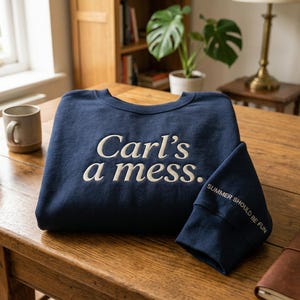 May include: Navy blue sweatshirt with the embroidered text "Carl's a mess." The sleeve cuff features the text "SUMMER SHOULD BE FUN." The sweatshirt is folded on a wooden table, with two mugs and a notebook visible.