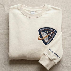 Puede incluir: Sudadera color crema con un parche azul marino con un cohete, la luna y el texto "RETURN TO THE MOON LUNAR FLYBY MISSION". El puño de la manga muestra el texto "FOR ALL HUMANITY" en azul marino.