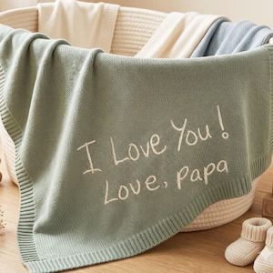 Op de afbeelding: Een saliegroene gebreide babydeken met de in het wit geborduurde tekst I Love You! Love, Papa. De deken is gedrapeerd over een geweven crèmekleurige mand, vergezeld door een klein houten berenfiguurtje en een paar crèmekleurige babyslofjes.