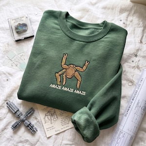May include: A forest green crewneck sweatshirt featuring an embroidered tan, multi-legged creature graphic centered on the chest. Below the creature, the text AMAZE AMAZE AMAZE is embroidered in white thread. The sweatshirt has long sleeves.