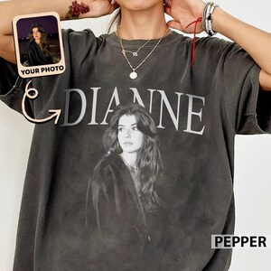 May include: Dark gray t-shirt featuring a black and white portrait of a person with the name "DIANNE" printed above. A small photo with the text "YOUR PHOTO" is in the upper left corner. The word "PEPPER" is in the lower right corner.