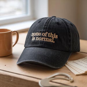 May include: Black baseball cap with the embroidered phrase "none of this is normal" in a light brown color. The cap is sitting on a wooden surface next to a mug and some papers.