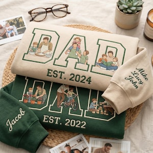 May include: Two sweatshirts and a sleeve are displayed. The sweatshirts are beige and dark green, embroidered with the word "DAD" and family illustrations. The beige sweatshirt says "EST. 2024". The green sweatshirt says "EST. 2022". The sleeve is beige and says "Mike Lucy John".
