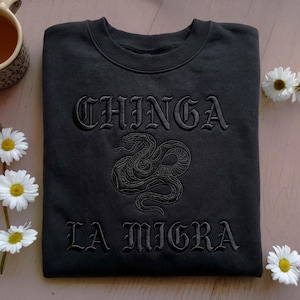 Puede incluir: Sudadera negra con la frase bordada "CHINGA LA MIGRA" en fuente gótica. Un diseño de serpiente bordada detallada está centrado debajo de la palabra "CHINGA". La sudadera se muestra sobre una superficie rosa claro con margaritas.