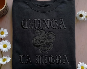 May include: Black sweatshirt with the embroidered text "CHINGA LA MIGRA" in a gothic font. A detailed embroidered snake design is centered below the word "CHINGA". The sweatshirt is displayed on a light pink surface with daisies.