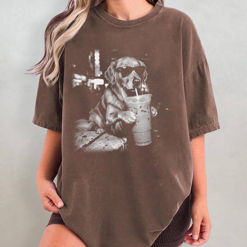 May include: Brown t-shirt featuring a grayscale graphic of a Golden Retriever wearing sunglasses and drinking from a clear iced coffee cup with a straw. The dog is sitting on a stone surface. The shirt has short sleeves.