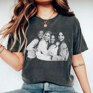 May include: Dark gray t-shirt featuring a black and white image of four smiling women embracing. The shirt has a crew neck and short sleeves. The image is centered on the front of the shirt.
