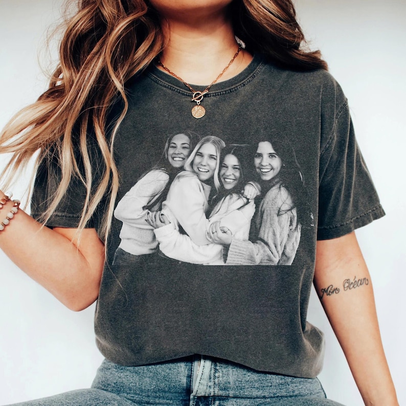 May include: Dark gray t-shirt featuring a black and white image of four smiling women embracing. The shirt has a crew neck and short sleeves. The image is centered on the front of the shirt.