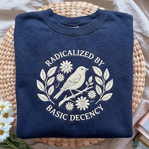 Peut inclure: Sweat-shirt bleu marine avec la phrase brodée "RADICALIZED BY BASIC DECENCY" entourant un oiseau sur une branche, orné de fleurs et de feuilles. Le sweat-shirt est plié sur un support tressé.