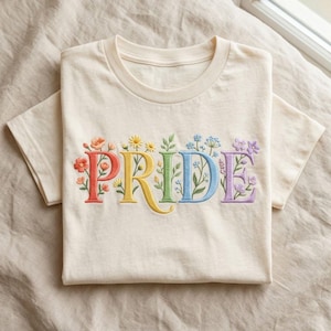 May include: Cream-colored t-shirt with the word "PRIDE" embroidered in colorful letters. Each letter is adorned with floral designs in shades of orange, yellow, green, blue, and purple. The shirt is folded neatly on a neutral-toned surface.