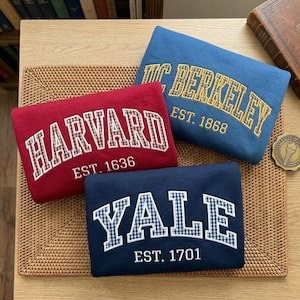 May include: Three folded sweatshirts in red, blue, and navy blue. The red sweatshirt features white text reading HARVARD EST. 1636. The blue one displays yellow text reading UC BERKELEY EST. 1868. The navy one shows white text reading YALE EST. 1701.