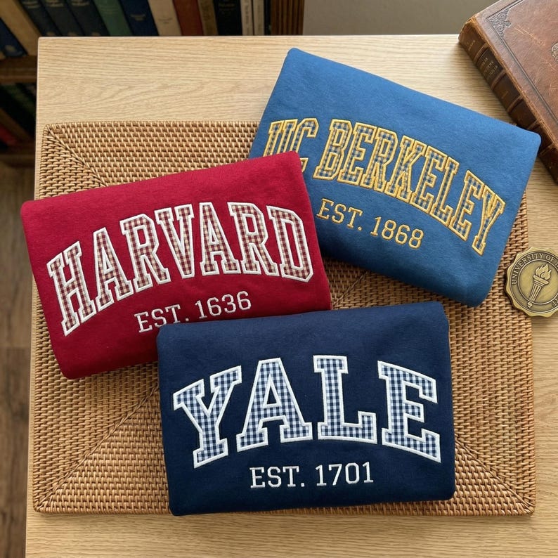 May include: Three folded sweatshirts in red, blue, and navy blue. The red sweatshirt features white text reading HARVARD EST. 1636. The blue one displays yellow text reading UC BERKELEY EST. 1868. The navy one shows white text reading YALE EST. 1701.