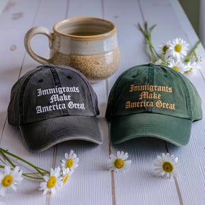 May include: Two baseball caps, one dark gray and one green, with the embroidered text "Immigrants Make America Great." A ceramic mug and daisies are also in the image.