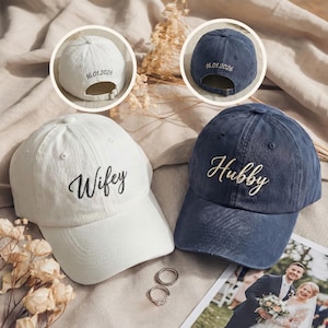 Puede incluir: Dos gorras de béisbol, una blanca y otra azul marino, con texto bordado. La gorra blanca dice "Wifey" y la gorra azul dice "Hubby". Ambas gorras tienen una fecha bordada en la parte trasera. Tema de boda.