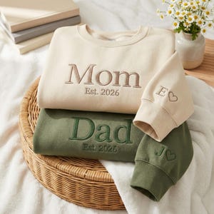 Puede incluir: Sudaderas crema y verde oliva bordadas con "Mom" y "Dad" y el año "Est. 2026". Las mangas están bordadas con iniciales y un corazón. Las sudaderas están dobladas en una cesta de mimbre.