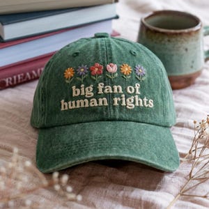 May include: A green baseball cap featuring embroidered flowers and the text "big fan of human rights" in white. The cap has a curved peak and an adjustable fabric strap. The flowers are in shades of orange, purple, pink, and yellow.
