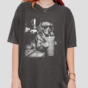 May include: Dark gray t-shirt featuring a black and white image of a Golden Retriever wearing sunglasses and holding an iced coffee. The dog is sitting on a stone surface. The shirt is a relaxed fit.