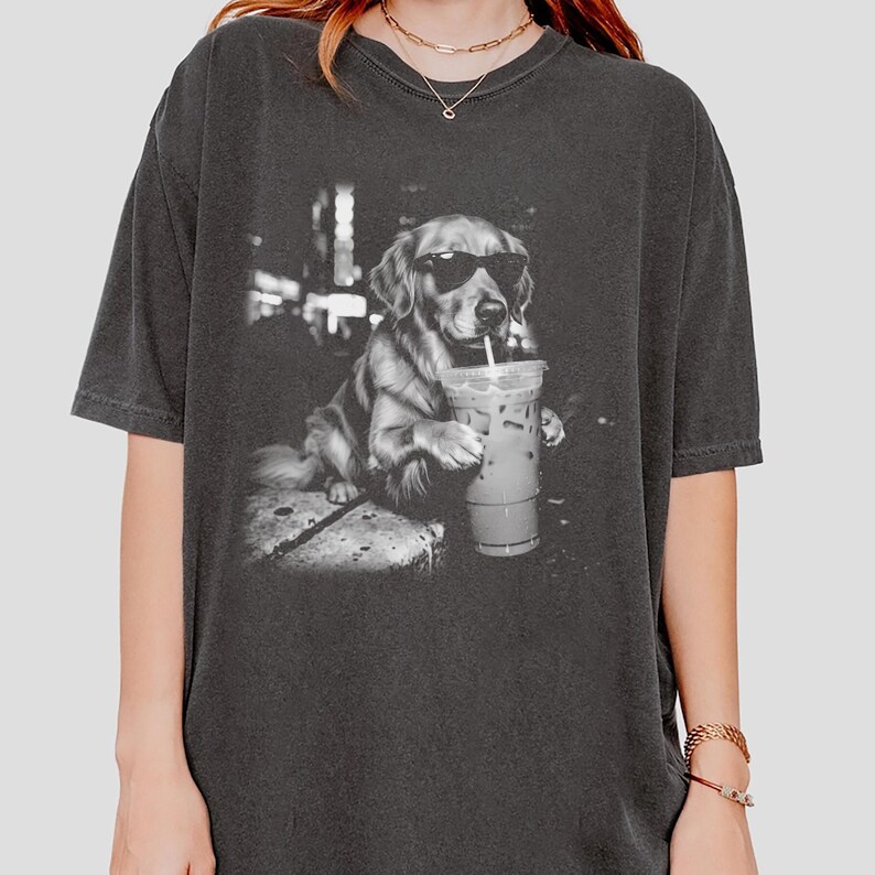 May include: Dark gray t-shirt featuring a black and white image of a Golden Retriever wearing sunglasses and holding an iced coffee. The dog is sitting on a stone surface. The shirt is a relaxed fit.