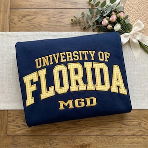 May include: A navy blue sweatshirt featuring the text UNIVERSITY OF FLORIDA in a yellow and white gingham pattern with a yellow border, and the letters MGD in yellow below. The sweatshirt is folded and displayed on a wooden surface.