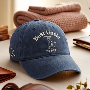 May include: A navy blue baseball cap with the text "Best Uncle BY PAR" in white. The cap features a golf-themed graphic and a crossed golf club design on the side. The hat is made of a soft, durable material.