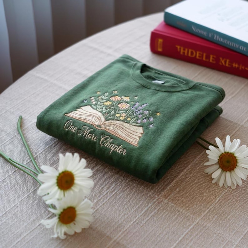 May include: Folded green sweatshirt with an embroidered design of an open book and flowers. The text "One More Chapter" is embroidered below the book. Two books are in the background, and daisies are in the foreground.
