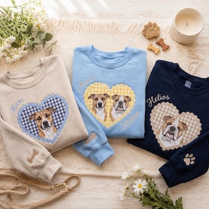 May include: Three crewneck sweatshirts in beige, light blue, and navy blue. Each features an embroidered heart with a gingham pattern containing dog portraits and names. The sleeves include embroidered bone or paw print details.
