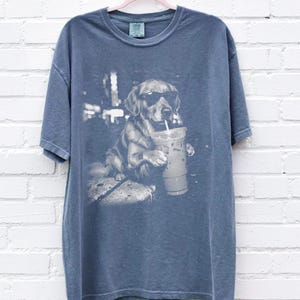 May include: Blue t-shirt featuring a black and white graphic of a golden retriever wearing sunglasses and holding a beverage with a straw. The dog is sitting and appears relaxed. The shirt has a crew neck and short sleeves.