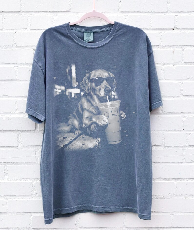 May include: Blue t-shirt featuring a black and white graphic of a golden retriever wearing sunglasses and holding a beverage with a straw. The dog is sitting and appears relaxed. The shirt has a crew neck and short sleeves.