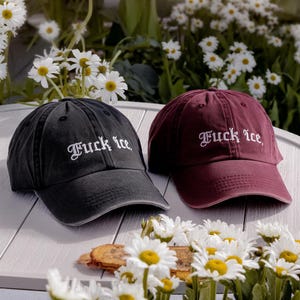 Puede incluir: Se muestran dos gorras de béisbol, una negra y otra burdeos. Cada gorra tiene la frase "Fuck Ice" bordada en blanco. Las gorras están colocadas sobre una superficie blanca, rodeadas de margaritas.