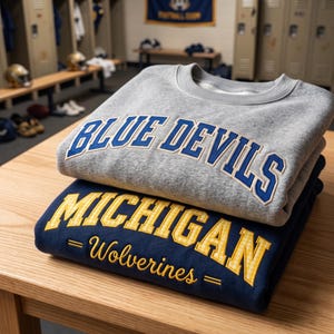 May include: A grey crewneck sweatshirt with blue embroidered text reading BLUE DEVILS stacked on a navy blue sweatshirt with yellow embroidered text reading MICHIGAN WOLVERINES. Both garments feature long sleeves and ribbed necklines.
