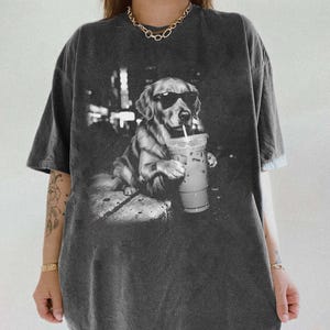 May include: Dark gray t-shirt featuring a black and white graphic of a golden retriever wearing sunglasses and holding an iced coffee. The dog is sitting on a stone surface. The shirt has short sleeves.