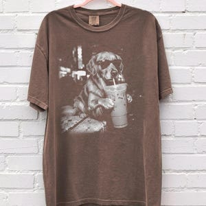 May include: Brown t-shirt featuring a black and white graphic of a Golden Retriever wearing sunglasses and drinking from a clear iced coffee cup with a straw. The dog is sitting on a stone surface.