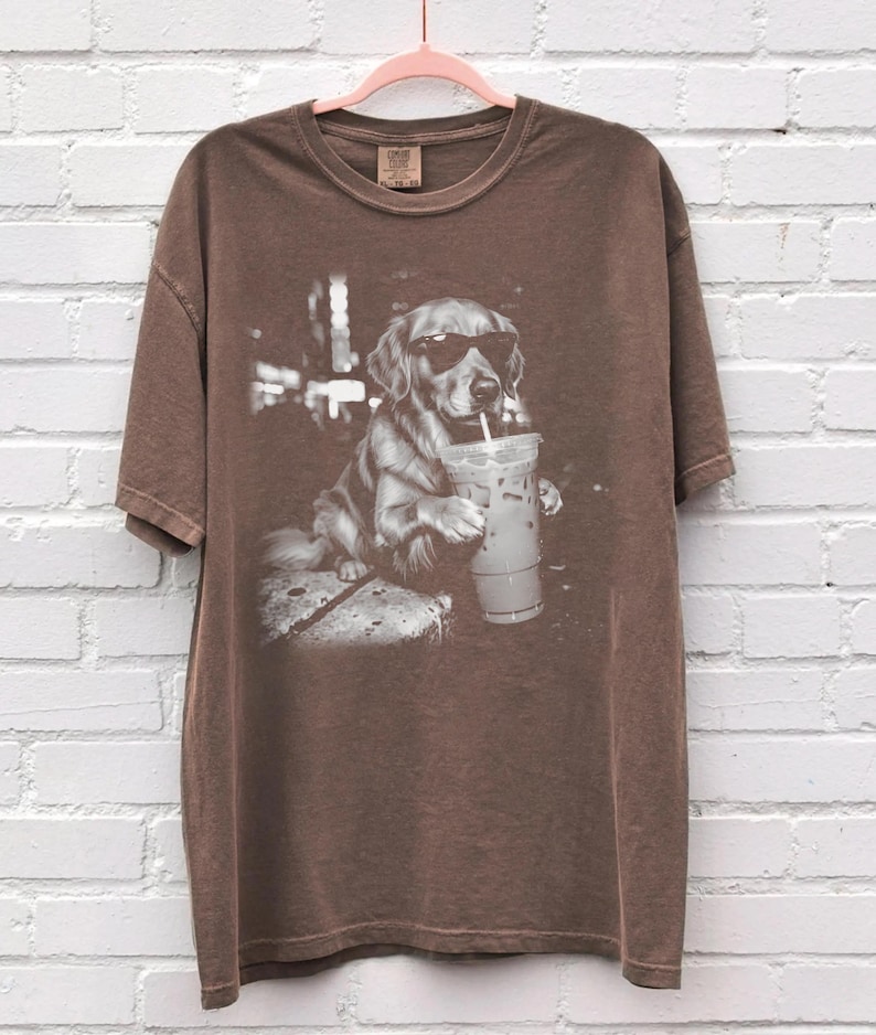 May include: Brown t-shirt featuring a black and white graphic of a Golden Retriever wearing sunglasses and drinking from a clear iced coffee cup with a straw. The dog is sitting on a stone surface.