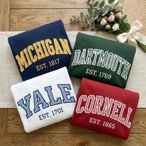 May include: Four sweatshirts in navy blue, green, white, and red. Each sweatshirt features a university name in a collegiate font with the establishment year. The sweatshirts are arranged on a neutral-toned surface.