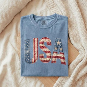 Op de afbeelding: Een lichtblauw T-shirt met het woord "USA" in een patchwork-ontwerp met de Amerikaanse vlag. Het shirt is netjes op een houten oppervlak gevouwen, met een crèmekleurige deken op de achtergrond.