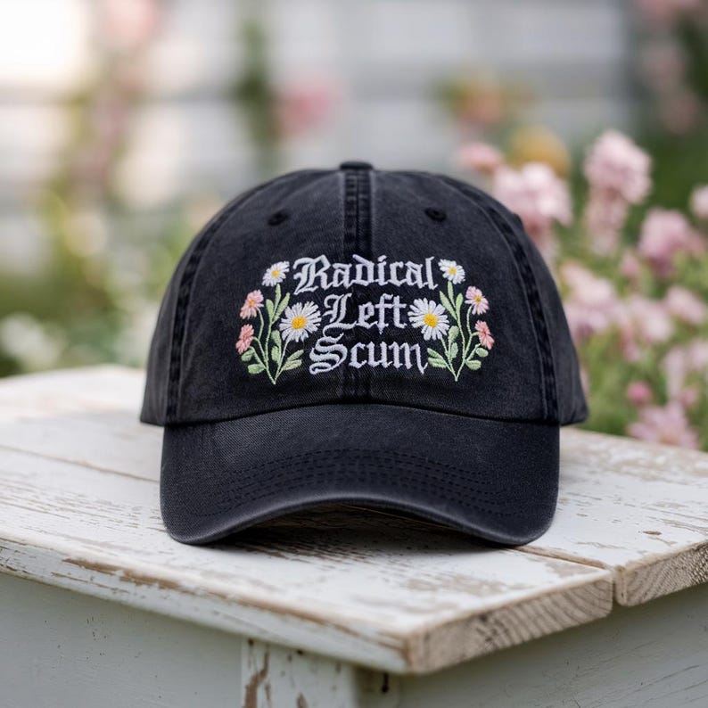 May include: Black baseball cap with the embroidered text "Radical Left Scum" surrounded by white daisies and pink flowers. The hat is sitting on a white wooden surface.