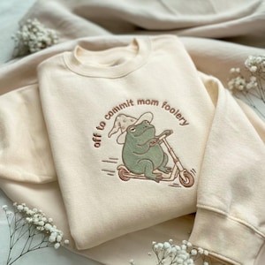 May include: Cream-colored sweatshirt with an embroidered design of a green frog wearing a witch hat and riding a scooter. The text "off to commit mom foolery" is arched around the frog. The sweatshirt is folded, with a soft, neutral background.