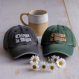 Può includere: Due cappellini da baseball, uno grigio scuro e uno verde, ricamati con "Chinga la Migra" in lettere bianche e dorate. Una tazza beige e margherite sullo sfondo. I cappellini sono un accessorio casual.