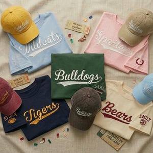 May include: Assortment of t-shirts and baseball caps in various colors, each featuring a team name in a script font. The shirts are light blue, pink, green, navy, and cream. The caps are yellow, tan, and brown. Some shirts have additional embroidered details.