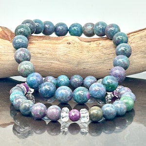 May include: Three bracelets made of blue, green, and pink gemstone beads. The bracelets are stacked on top of each other on a wooden surface.
