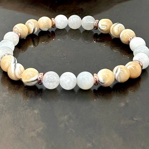 Puede incluir: Un brazalete hecho de cuentas blancas y beige con detalles dorados. Las cuentas son redondas y tienen un acabado liso y pulido.